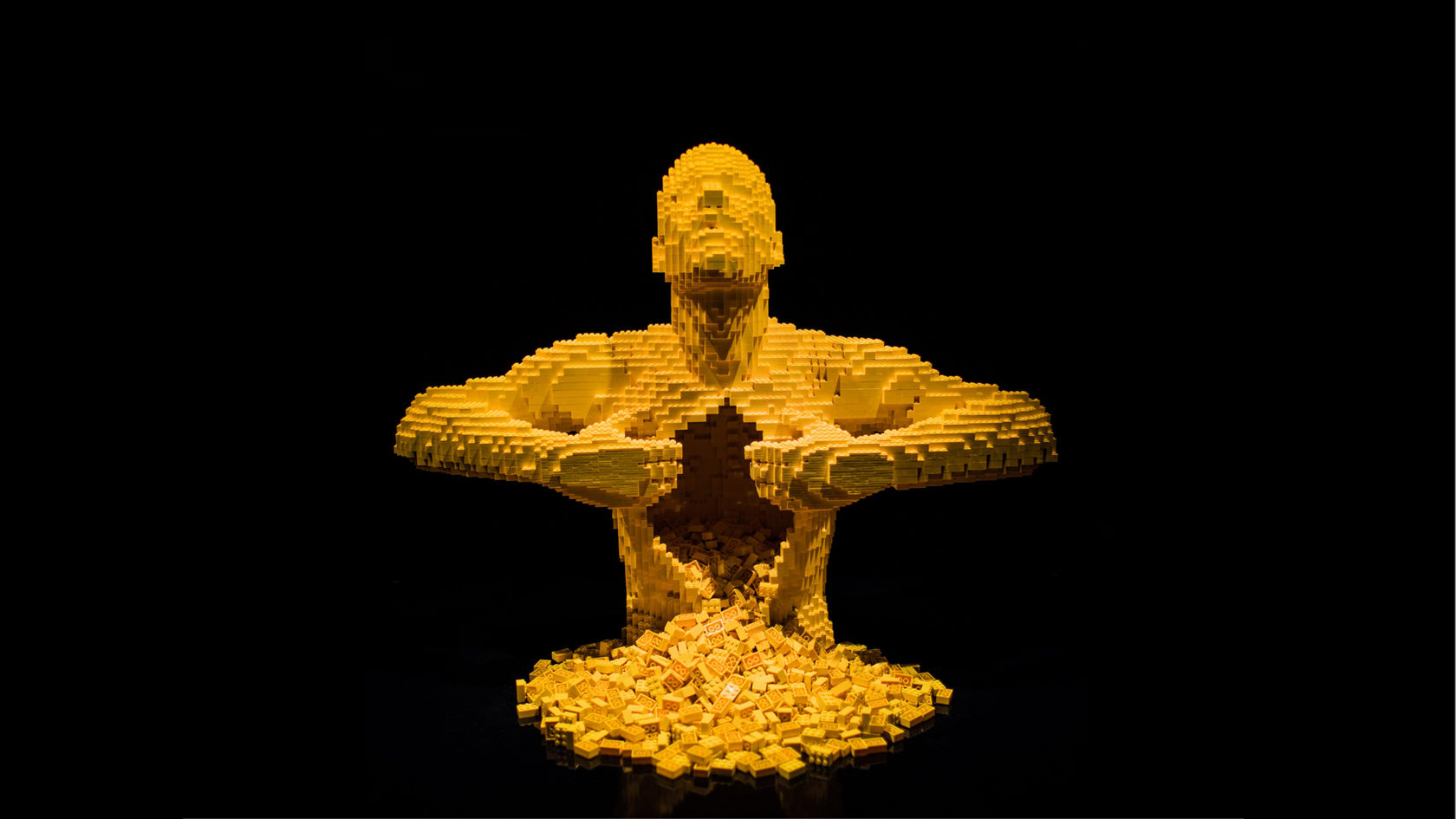 Скульптура из жёлтых кубиков LEGO на чёрном фоне: человек раскрывает руками грудь, и из неё высыпаются кубики